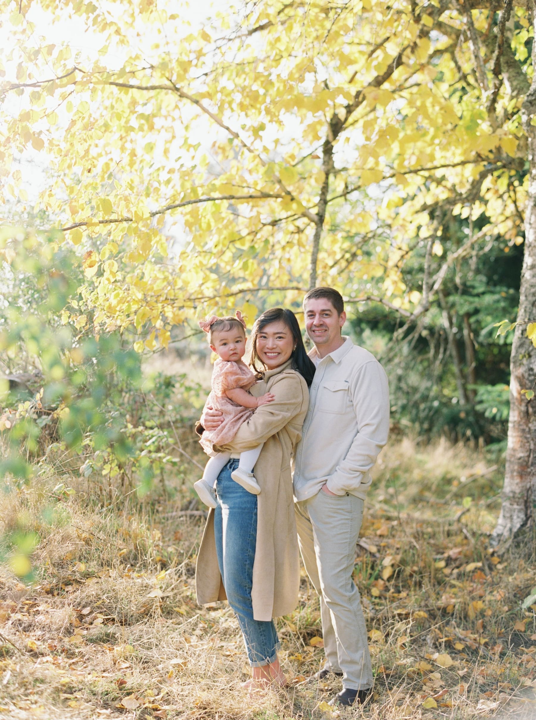 A woman holding a baby and a man stand together outdoors in a sunny, wooded area with yellow autumn leaves, perfect for a Kirkland family photographer capturing heartfelt moments.
