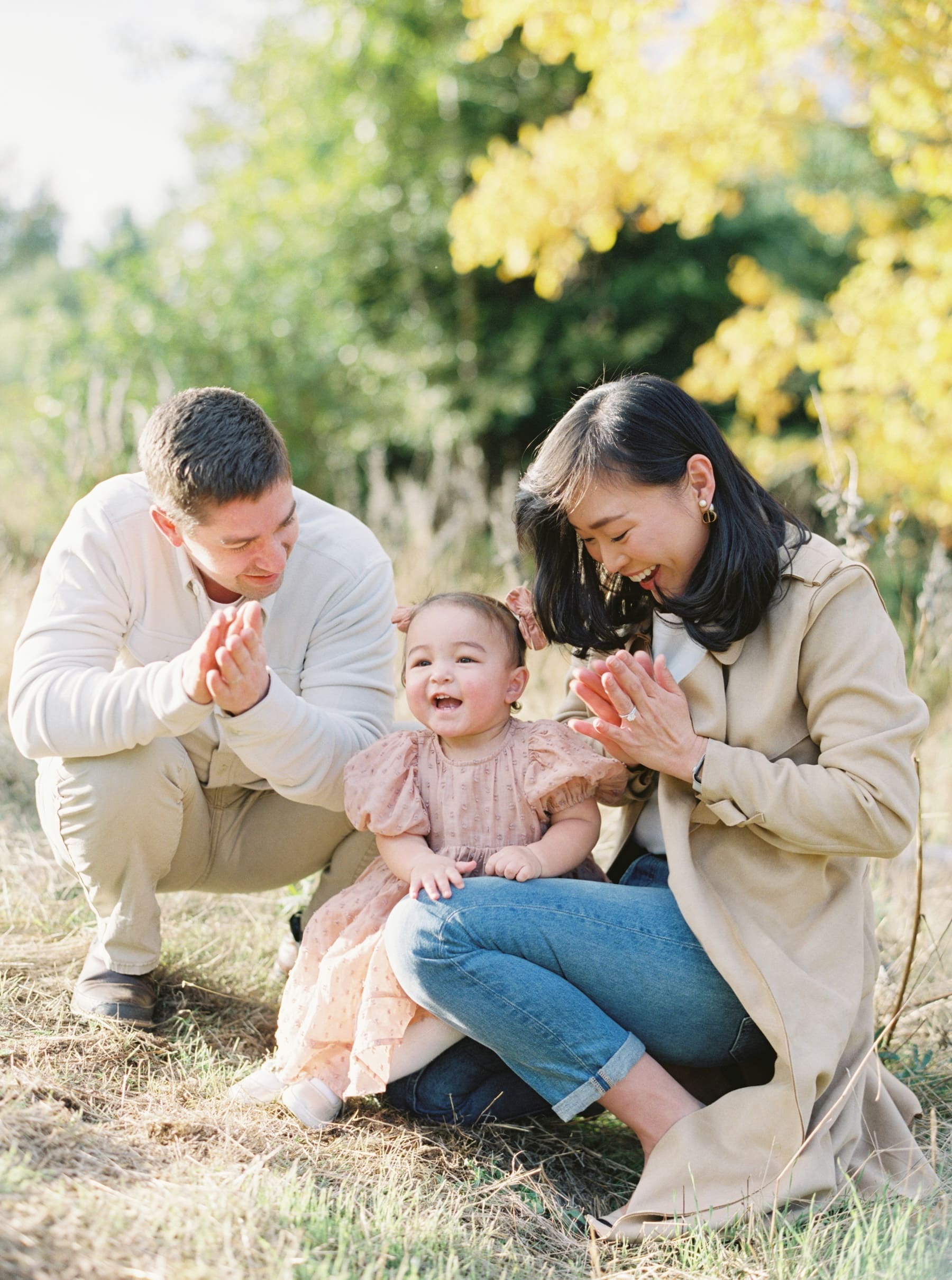 Kirkland family photography at Juanita Bay Park. Happy family clapping hands with baby girl in a pink dress outdoors.