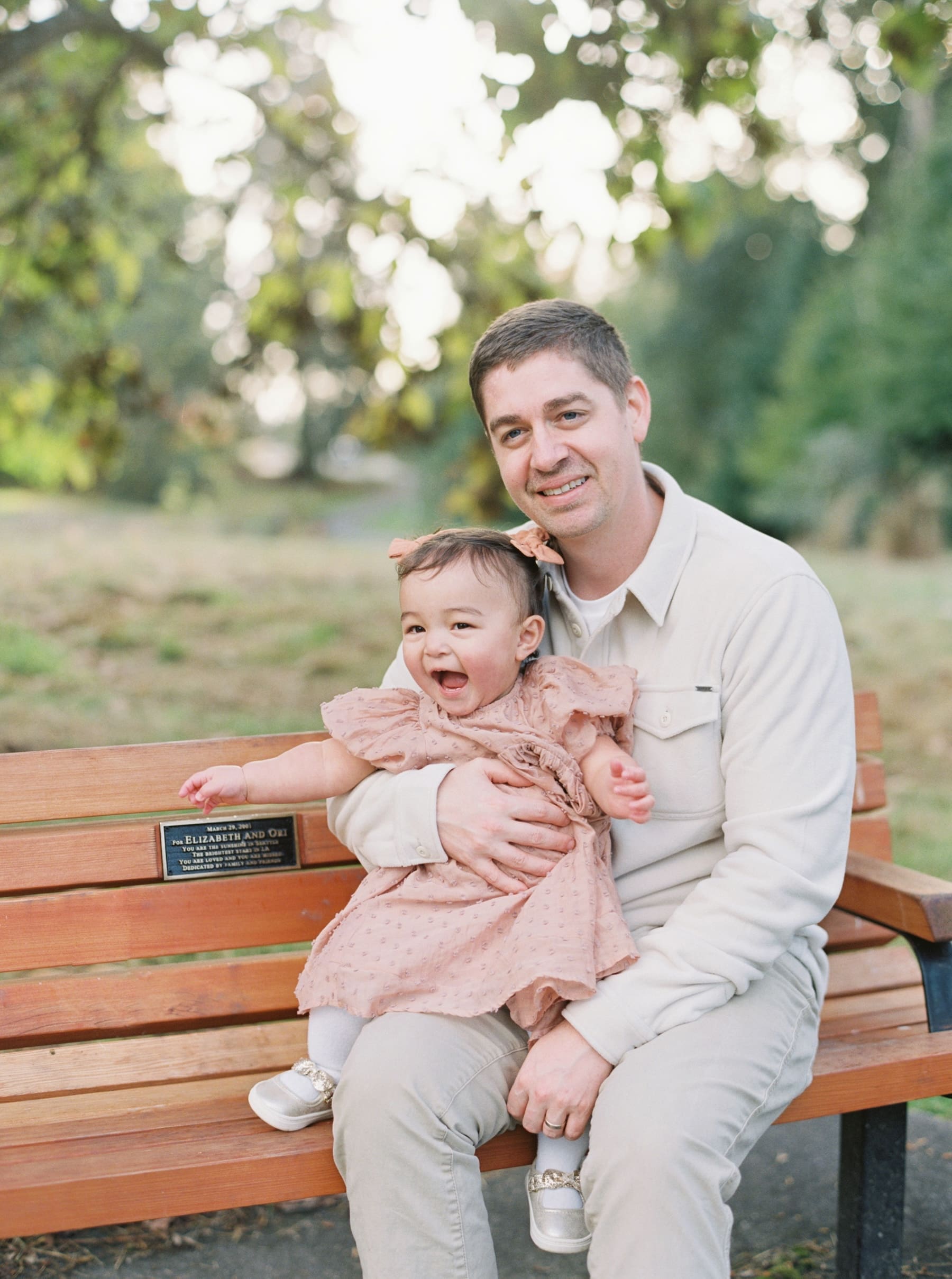 A man in a light shirt and pants sits on a park bench holding a smiling toddler girl in a pink dress and bow. The blurred greenery hints at Juanita Bay Park family photos by a Kirkland family photographer, capturing this pleasant outdoor moment.