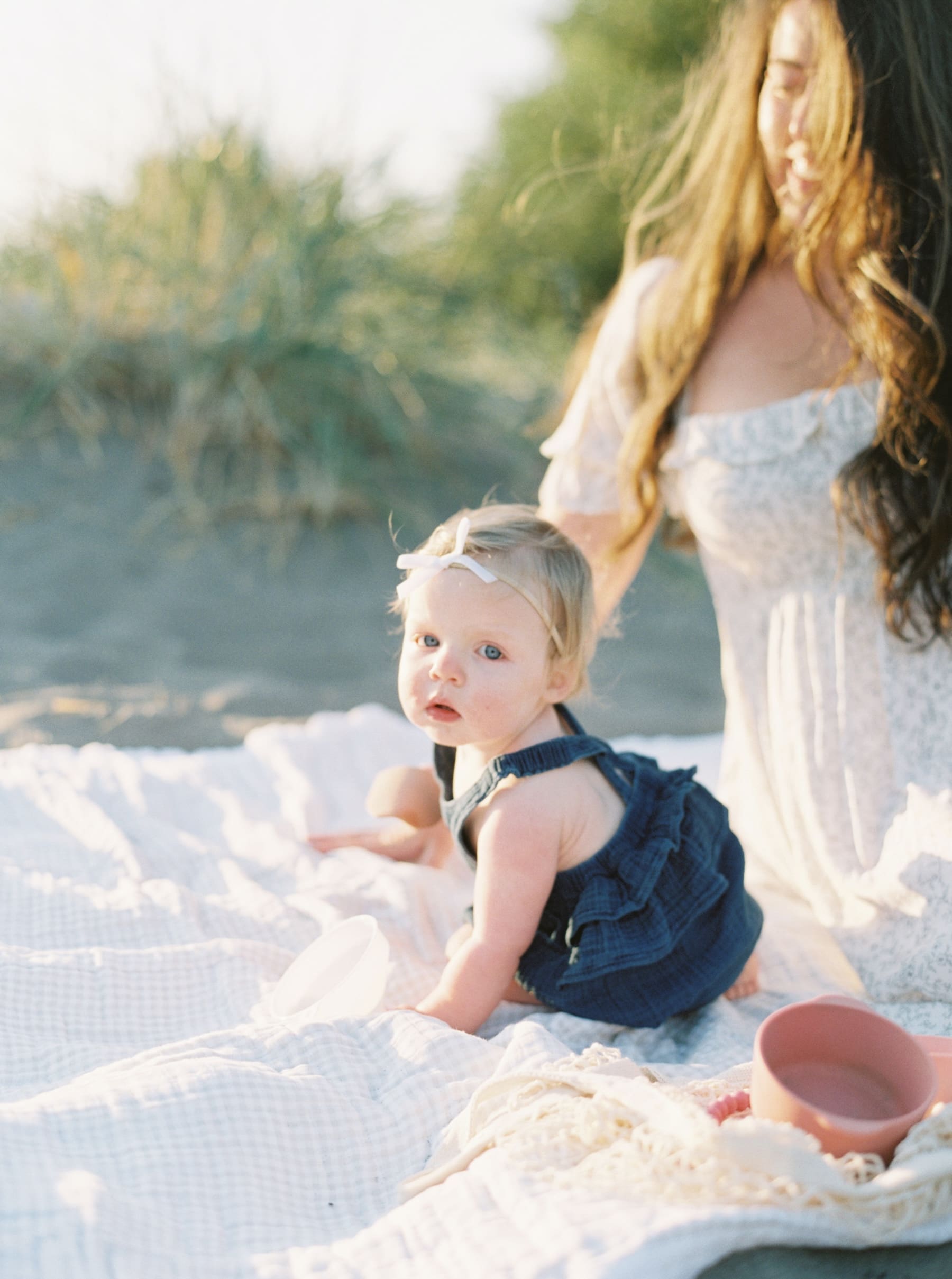 one year old baby in navy blue romper on sandy beach with mom on blanket