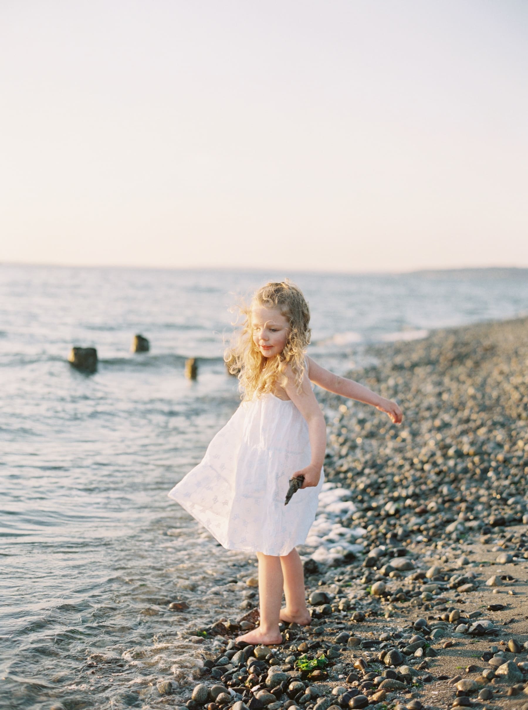 little girl in white dress at beach throwing rocks in the water