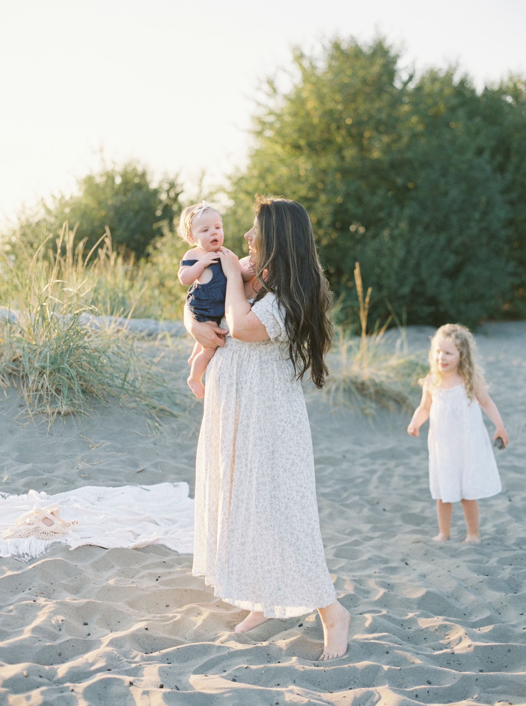 mom holding baby while wearing long white dress with younger daughter in the backgrounf