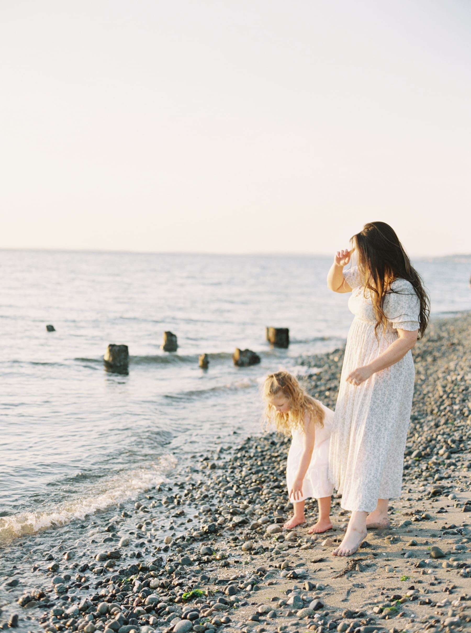 mom in long white dress at the waters edge on the beach playing with young daughter