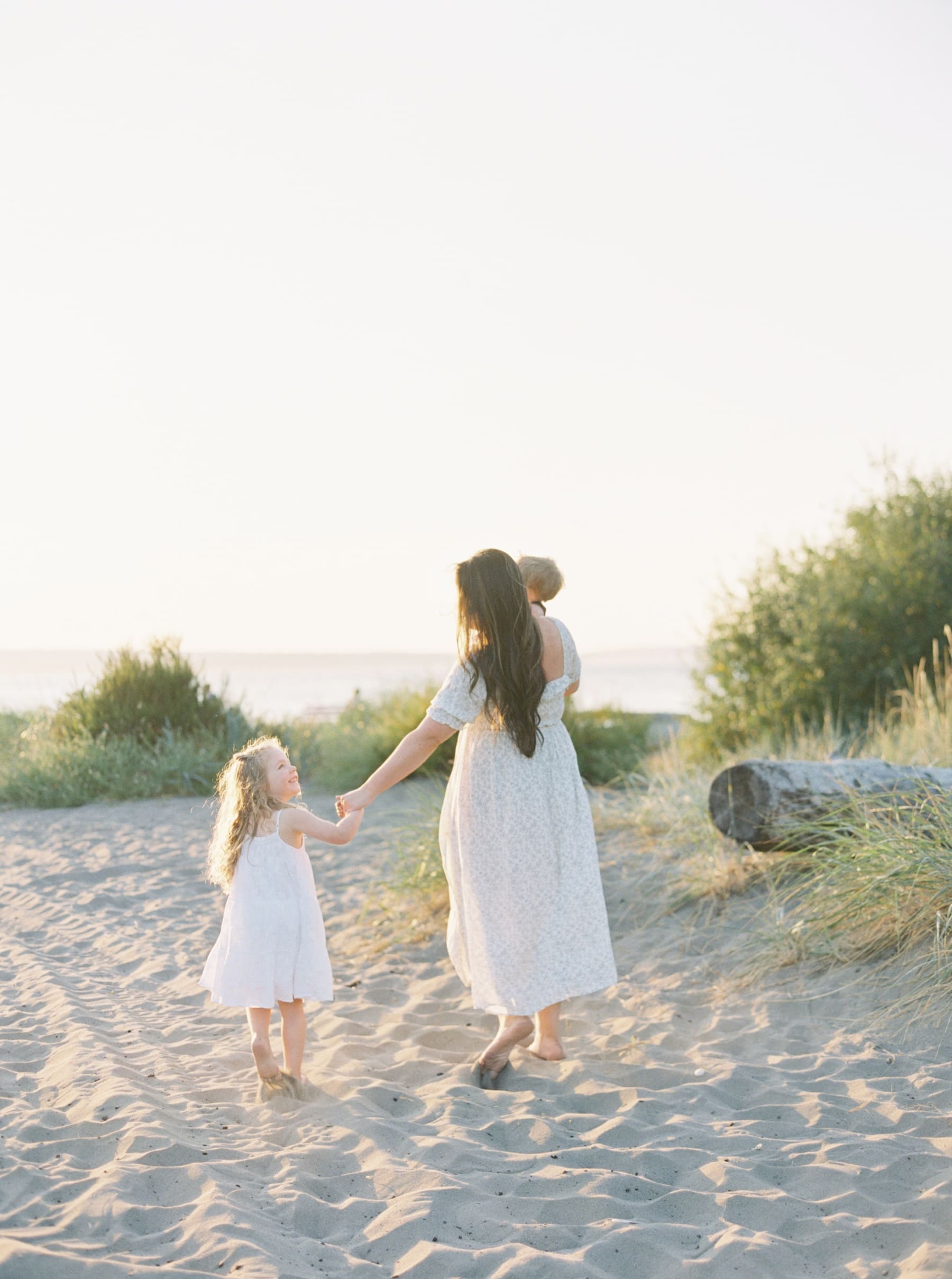 mother walking on sandy beach in white dress holding daughters hand and carrying baby