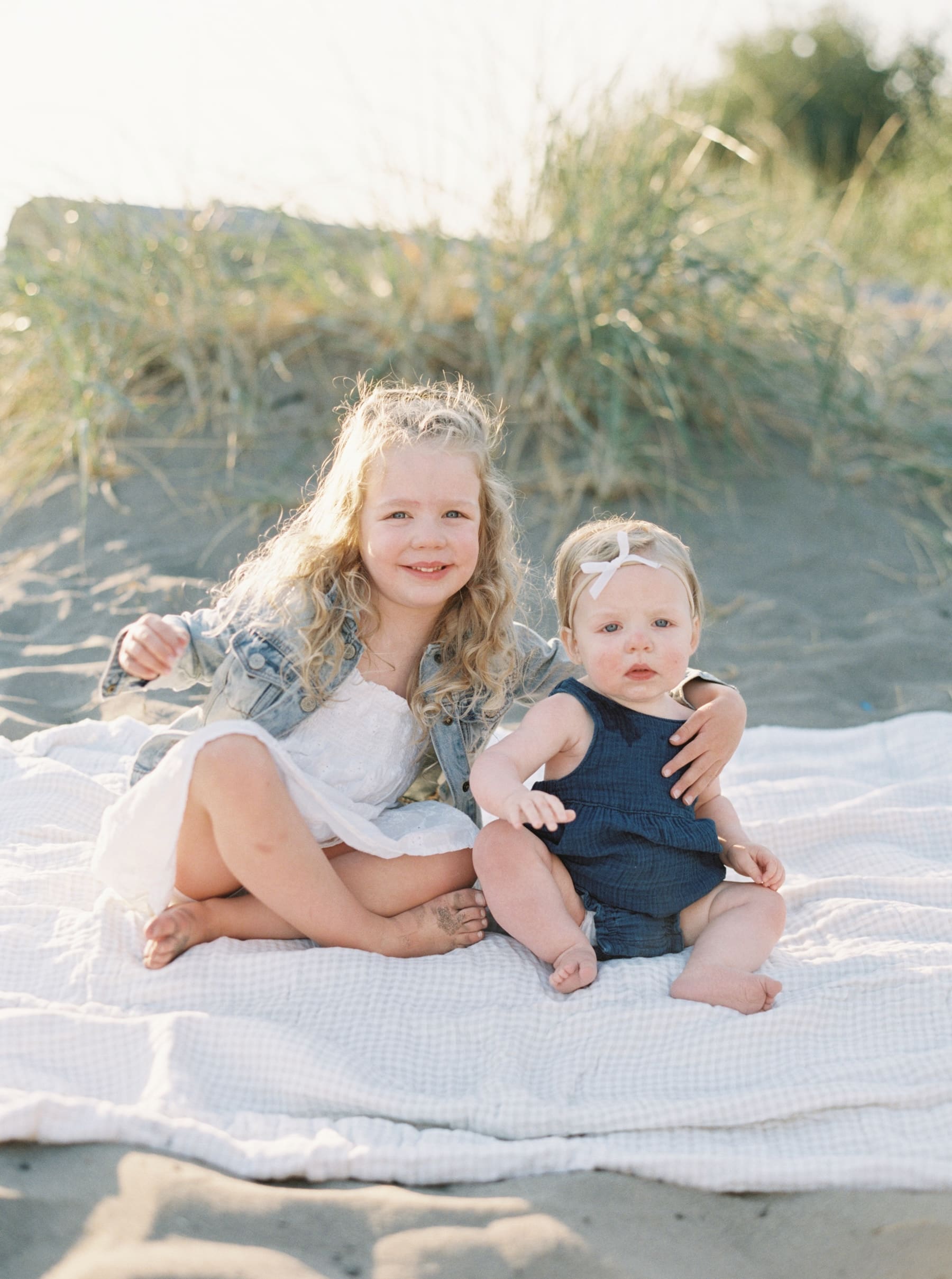 young girl and baby sister looking at the camera sitting on beach blanket