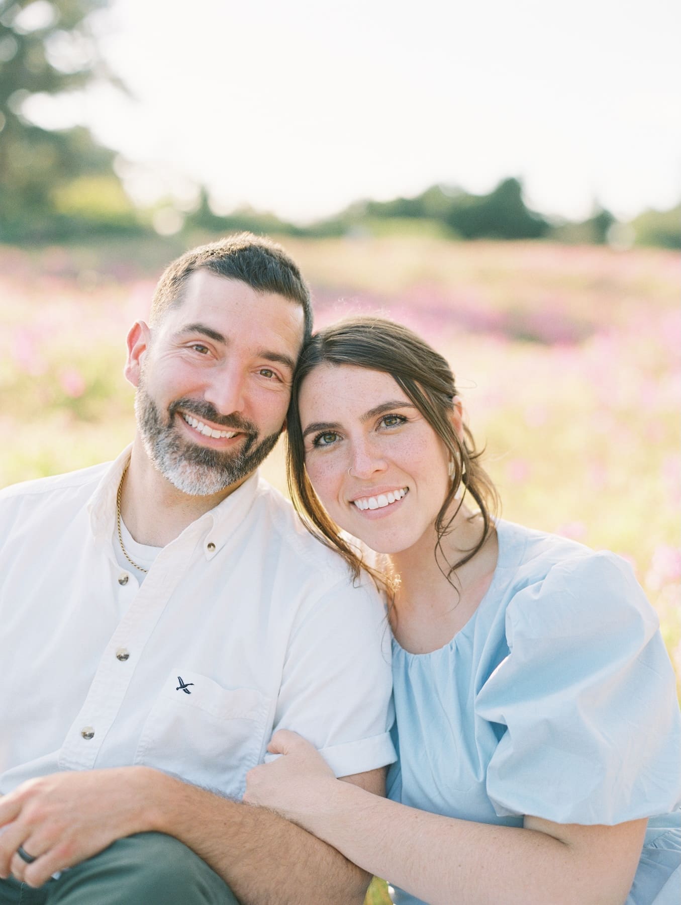 Man and woman leaning heads together smiling while sitting in a soft-focus field of pink flowers.