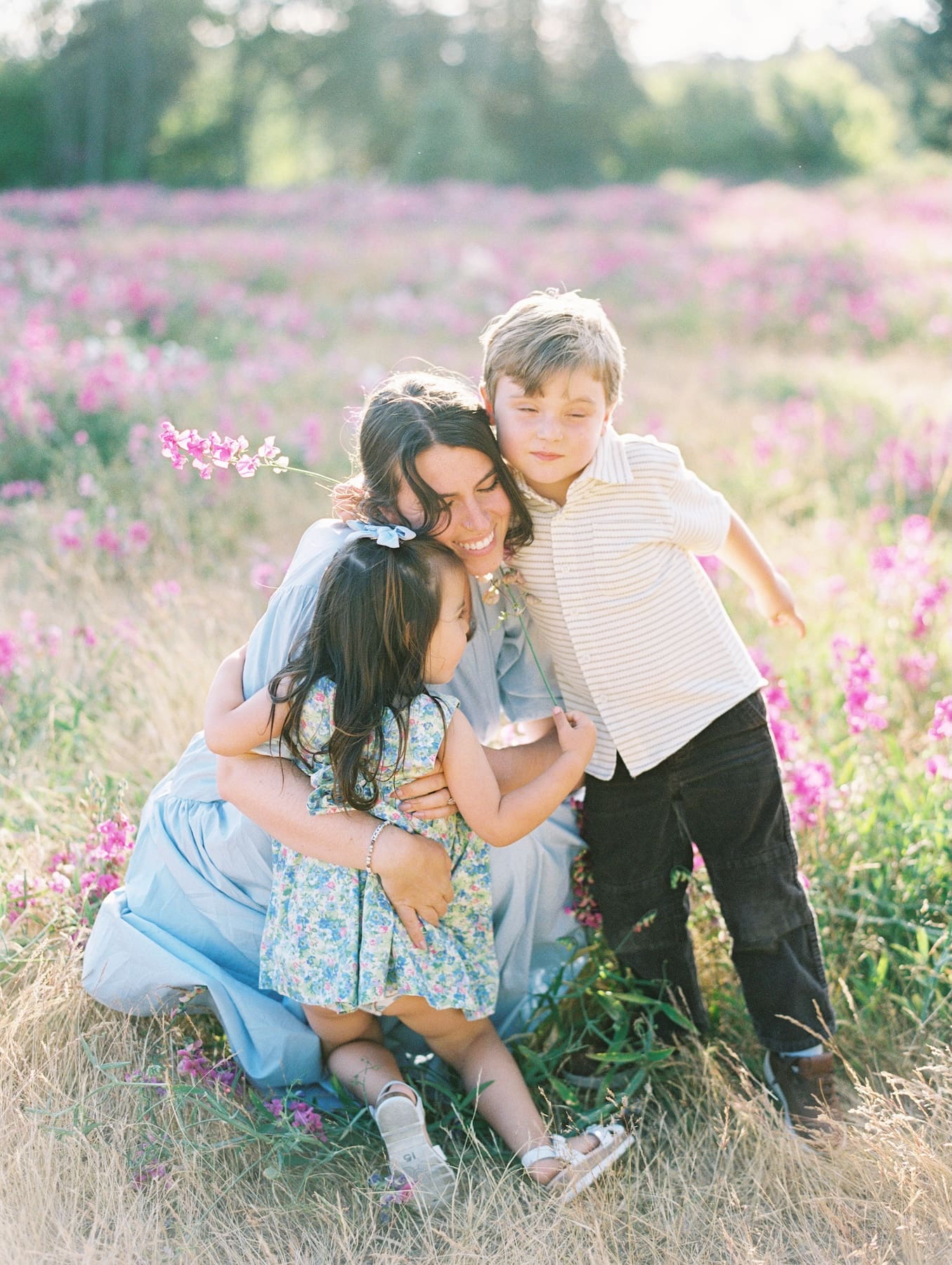 Mother kneeling in a meadow of pink flowers while embracing her young son and daughter.