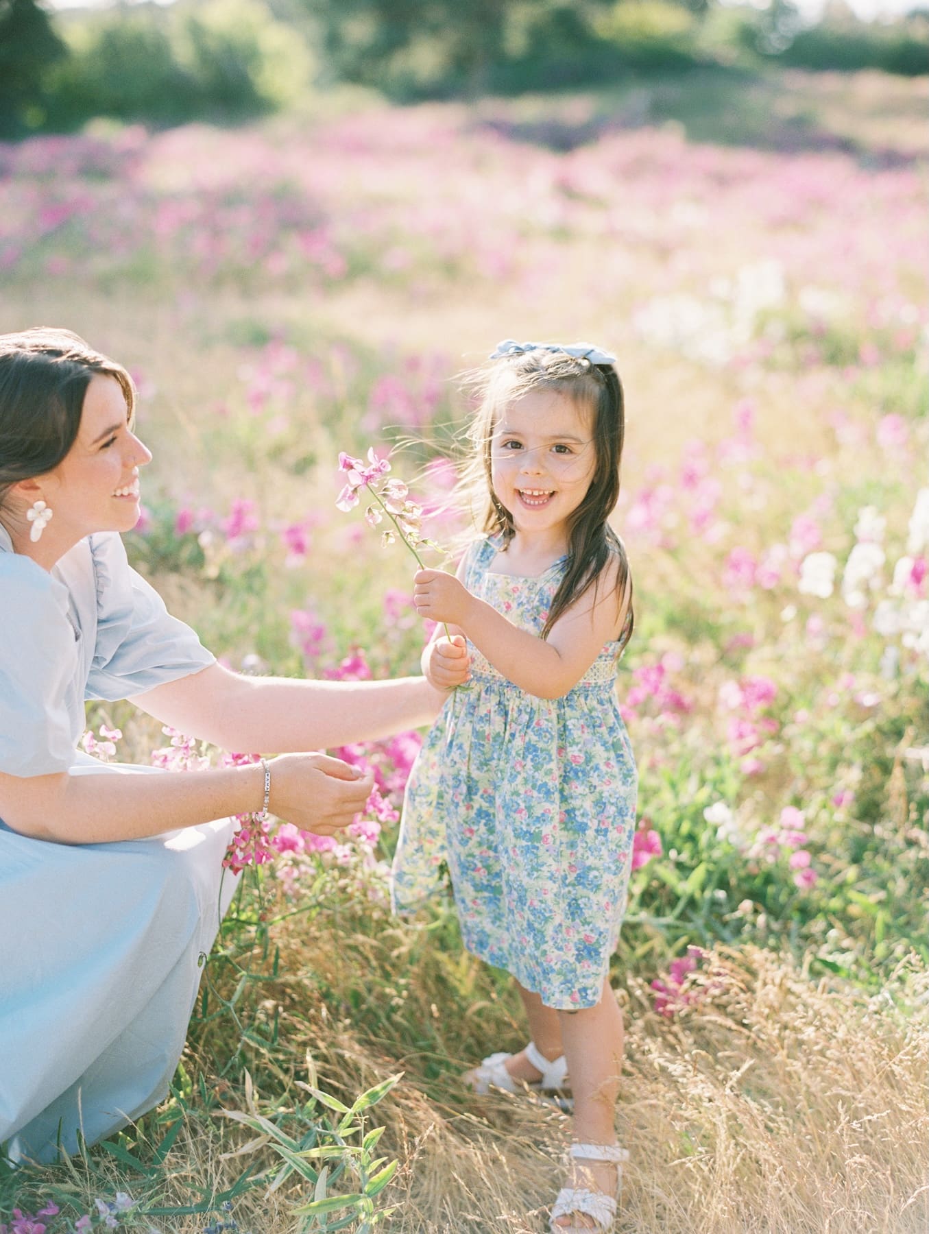 Young girl in a floral dress smiles while holding a small pink flower out to her mother in a sunny field.